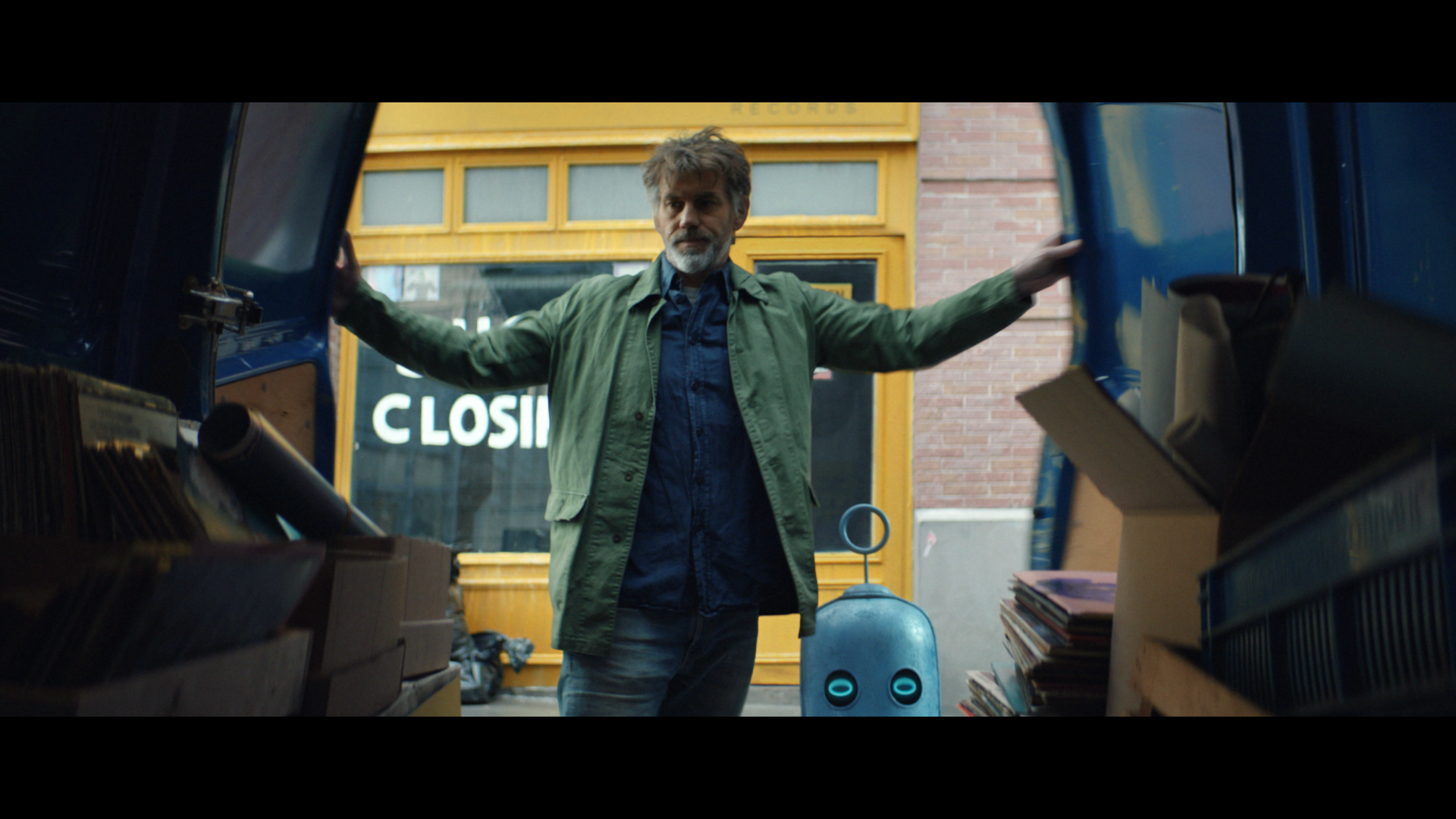 A man in a green shirt opening the back doors of a van with a robot next to him