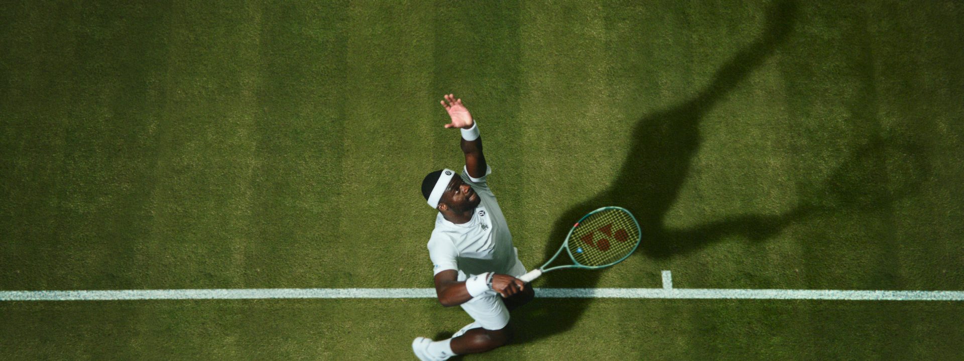 Frances Tiafoe prepares to serve during a tennis match on grass, captured from above with his shadow stretched across the court.