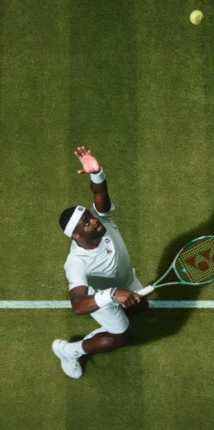 Frances Tiafoe prepares to serve during a tennis match on grass, captured from above with his shadow stretched across the court.