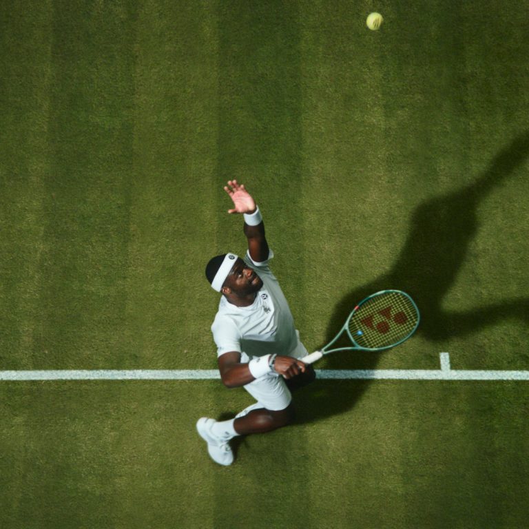 Frances Tiafoe prepares to serve during a tennis match on grass, captured from above with his shadow stretched across the court.
