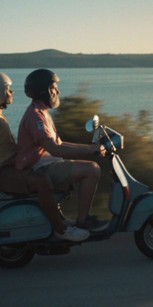Two older men ride a vintage blue moped along a coastal road at sunset, capturing the freedom of spontaneous travel.