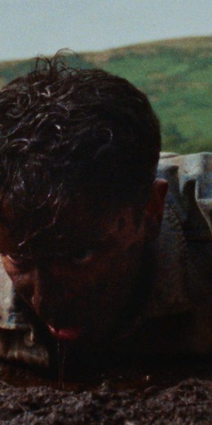 A man in a denim jacket lies face down in a muddy field, covered in dirt, struggling to push himself up, with green hills in the background.