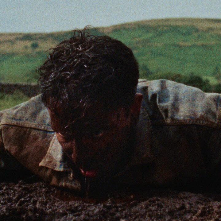 A man in a denim jacket lies face down in a muddy field, covered in dirt, struggling to push himself up, with green hills in the background.