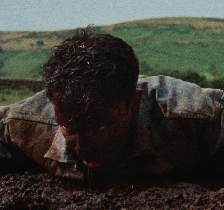 A man in a denim jacket lies face down in a muddy field, covered in dirt, struggling to push himself up, with green hills in the background.
