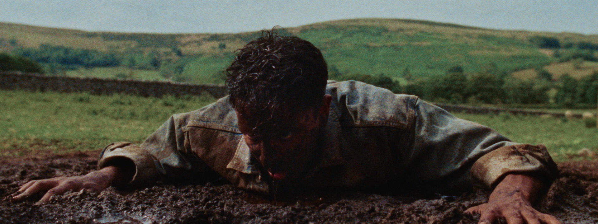 A man in a denim jacket lies face down in a muddy field, covered in dirt, struggling to push himself up, with green hills in the background.