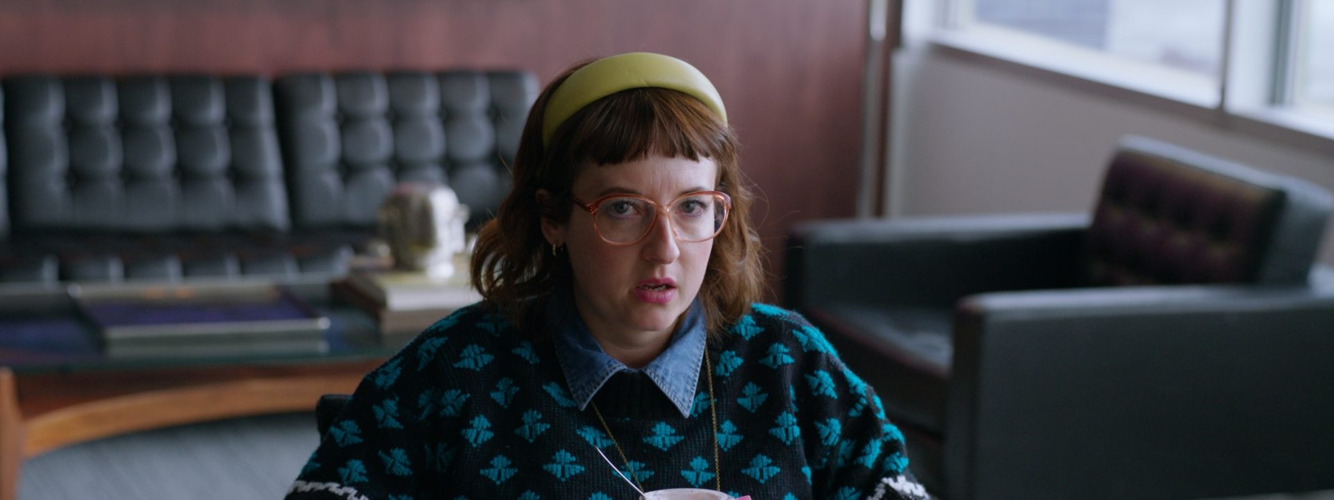 A woman wearing glasses, a cat patterned jumper, and a clock pendant sits in an office holding a yogurt, looking surprised.