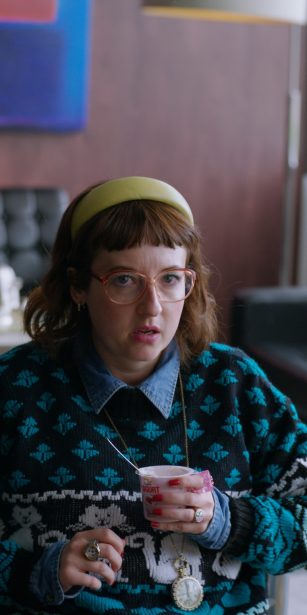 A woman wearing glasses, a cat patterned jumper, and a clock pendant sits in an office holding a yogurt, looking surprised.