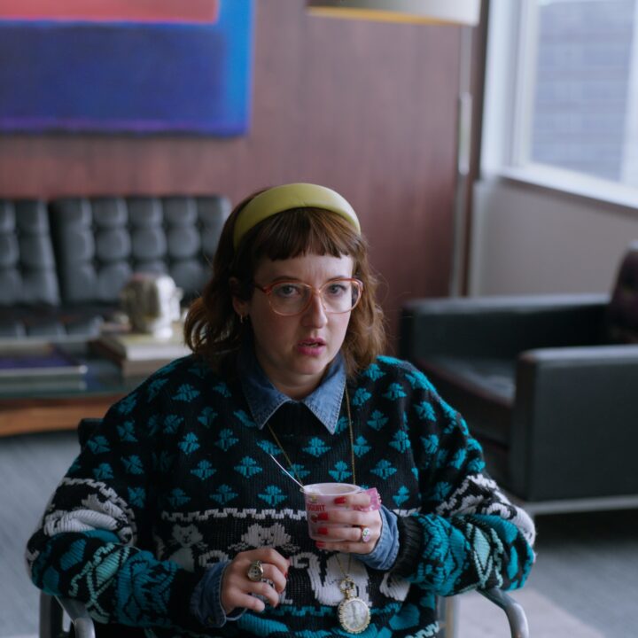 A woman wearing glasses, a cat patterned jumper, and a clock pendant sits in an office holding a yogurt, looking surprised.