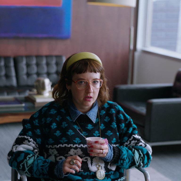 A woman wearing glasses, a cat patterned jumper, and a clock pendant sits in an office holding a yogurt, looking surprised.