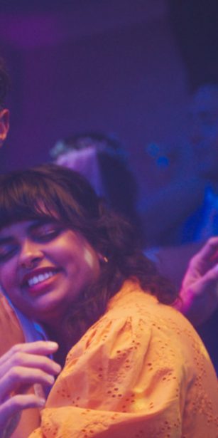 A vicar in white robes looks uncomfortable while slow dancing with a smiling woman in an orange dress at a party, under purple disco lights.