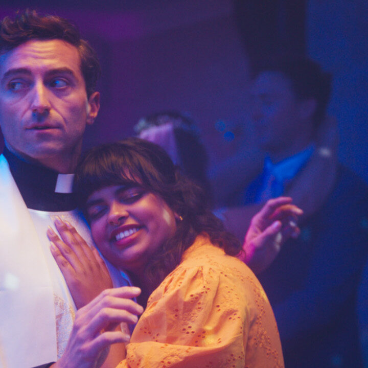 A vicar in white robes looks uncomfortable while slow dancing with a smiling woman in an orange dress at a party, under purple disco lights.