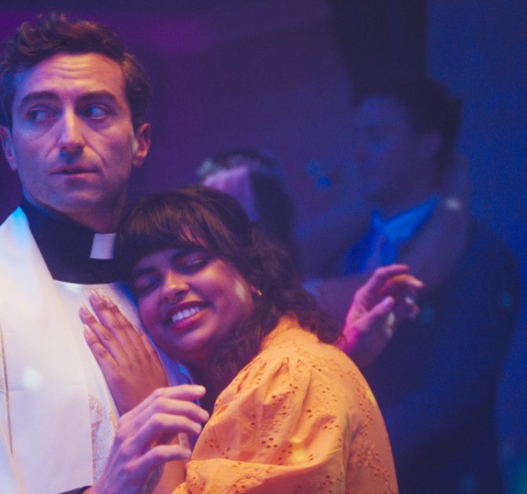 A vicar in white robes looks uncomfortable while slow dancing with a smiling woman in an orange dress at a party, under purple disco lights.