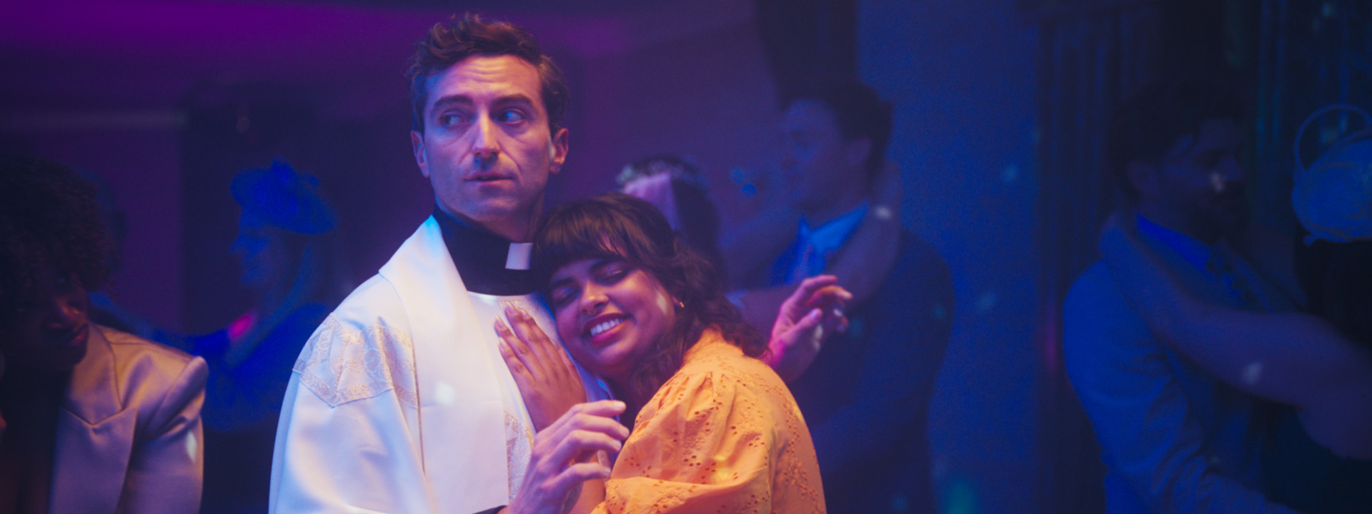 A vicar in white robes looks uncomfortable while slow dancing with a smiling woman in an orange dress at a party, under purple disco lights.