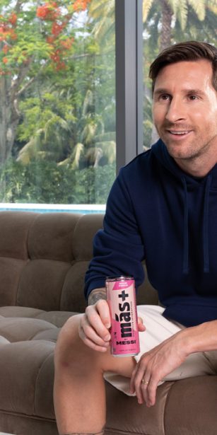 Lionel Messi seated on a sofa, holding a pink Más+ drink can, wearing a navy hoodie, with a tropical garden visible behind him.