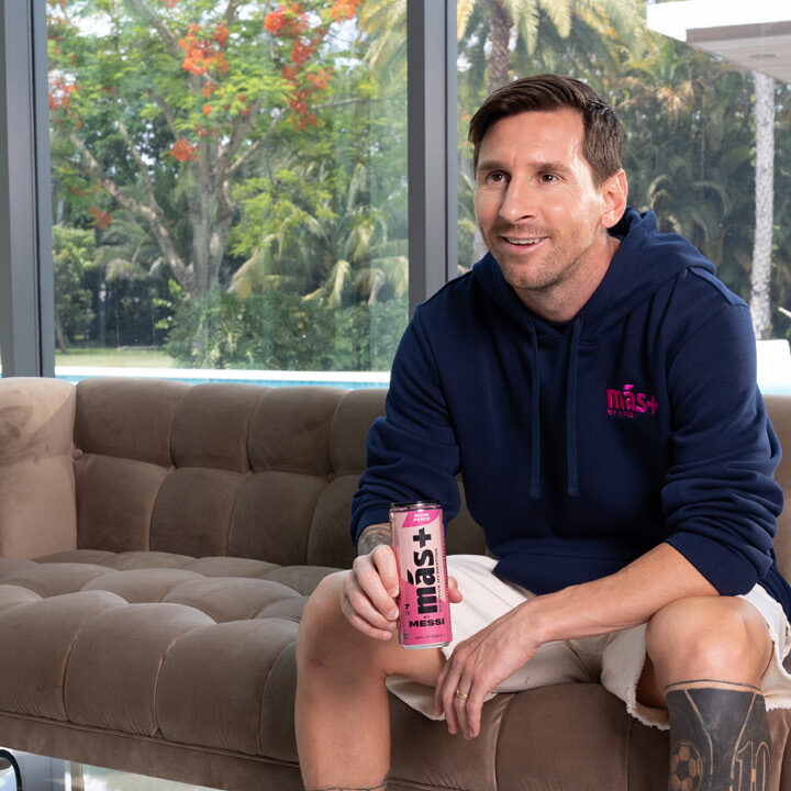 Lionel Messi seated on a sofa, holding a pink Más+ drink can, wearing a navy hoodie, with a tropical garden visible behind him.