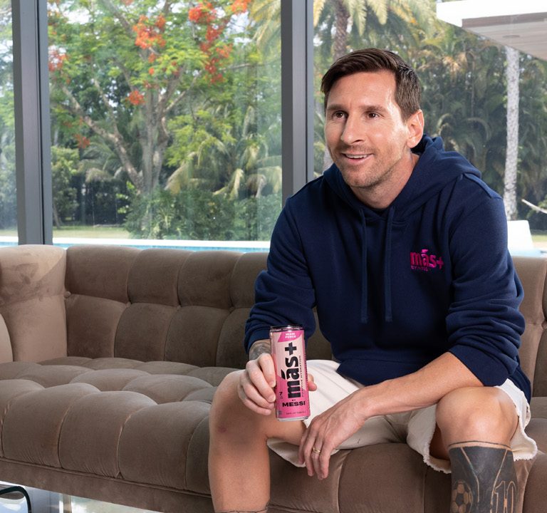 Lionel Messi seated on a sofa, holding a pink Más+ drink can, wearing a navy hoodie, with a tropical garden visible behind him.