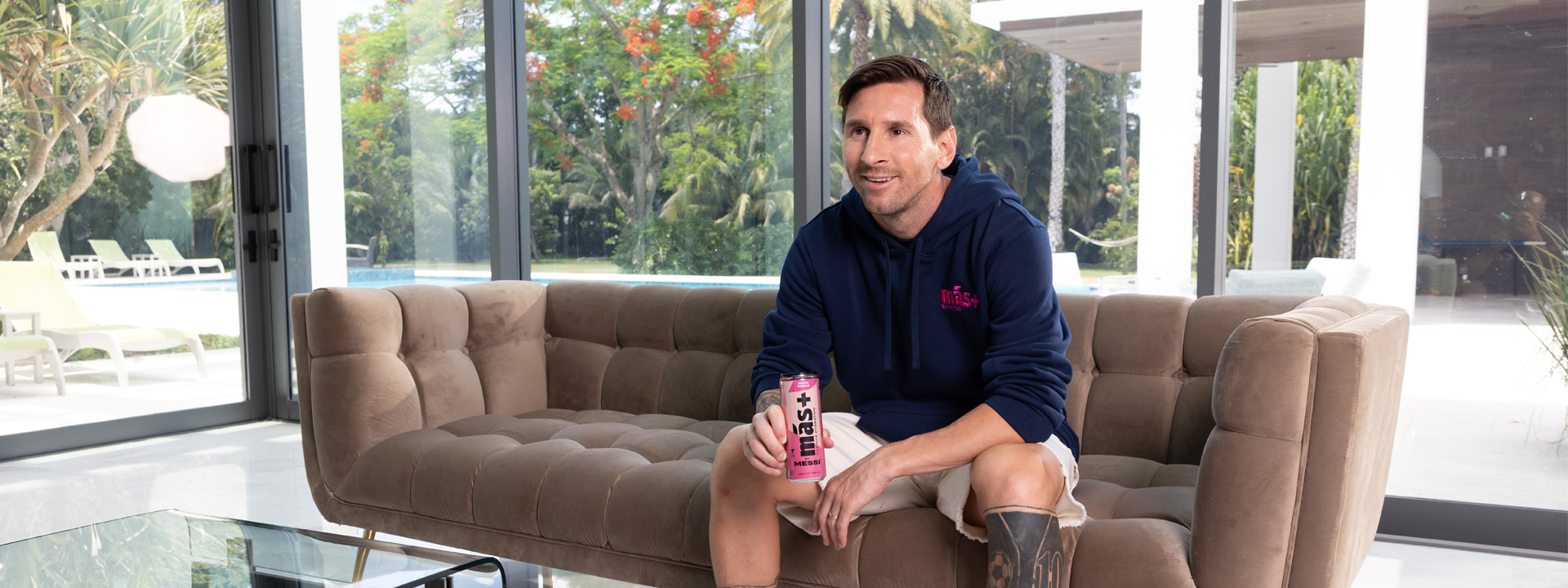 Lionel Messi seated on a sofa, holding a pink Más+ drink can, wearing a navy hoodie, with a tropical garden visible behind him.