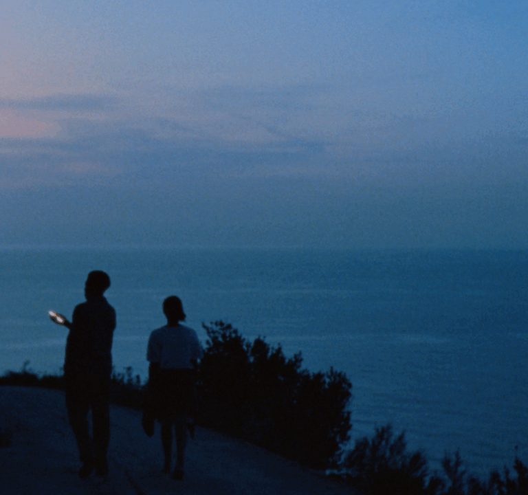 Two people on a coastal path at sunset illuminated by the light of a phone screen