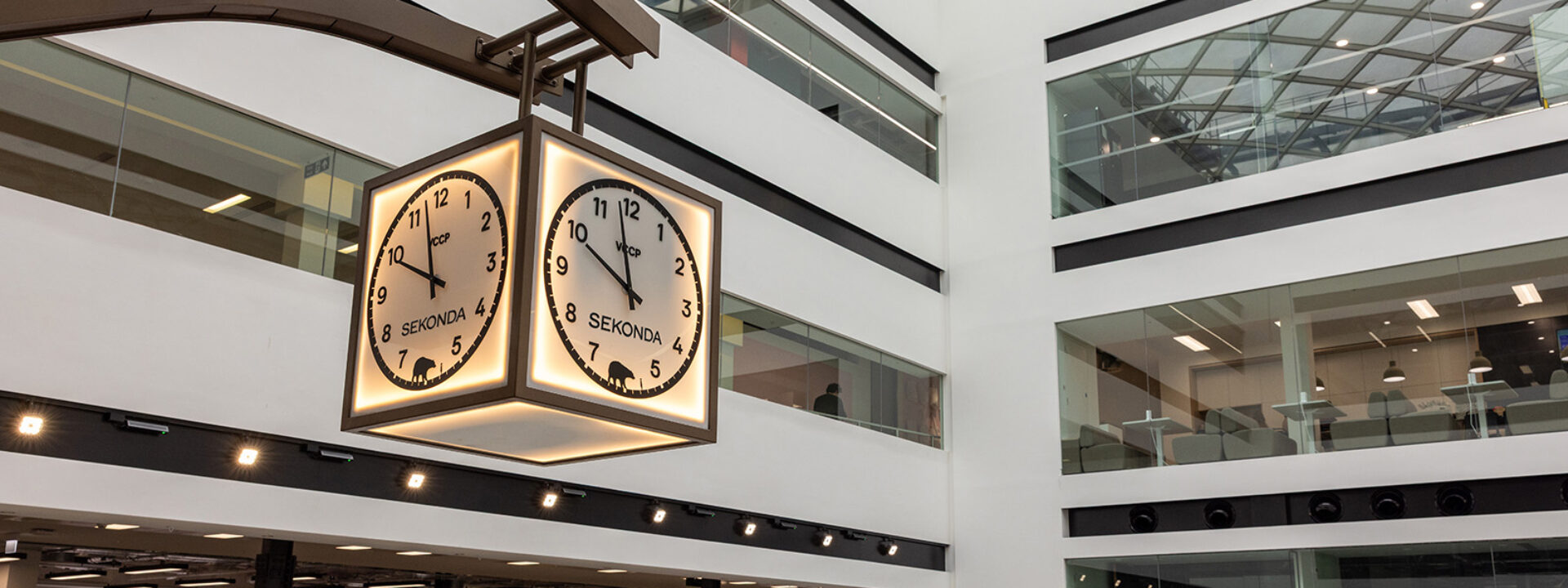 Custom VCCP and Sekonda branded hanging square clock suspended in a high-ceiling corporate office building, VCCP in London.