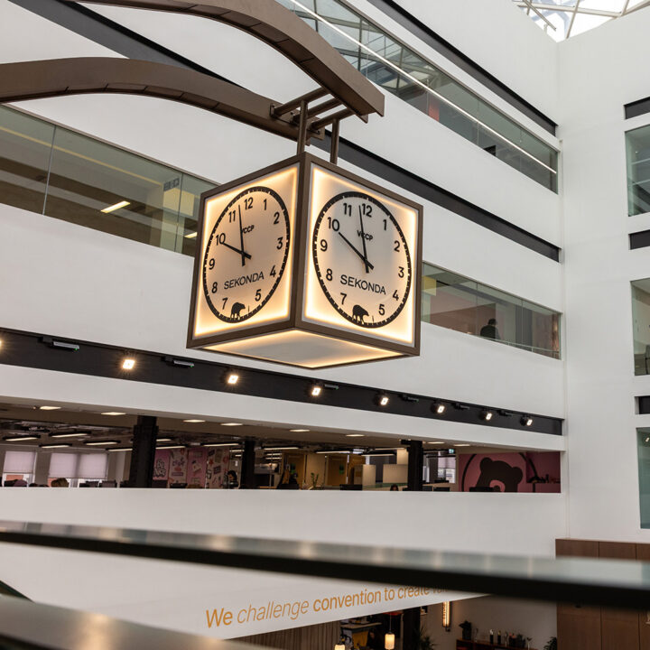 Custom VCCP and Sekonda branded hanging square clock suspended in a high-ceiling corporate office building, VCCP in London.