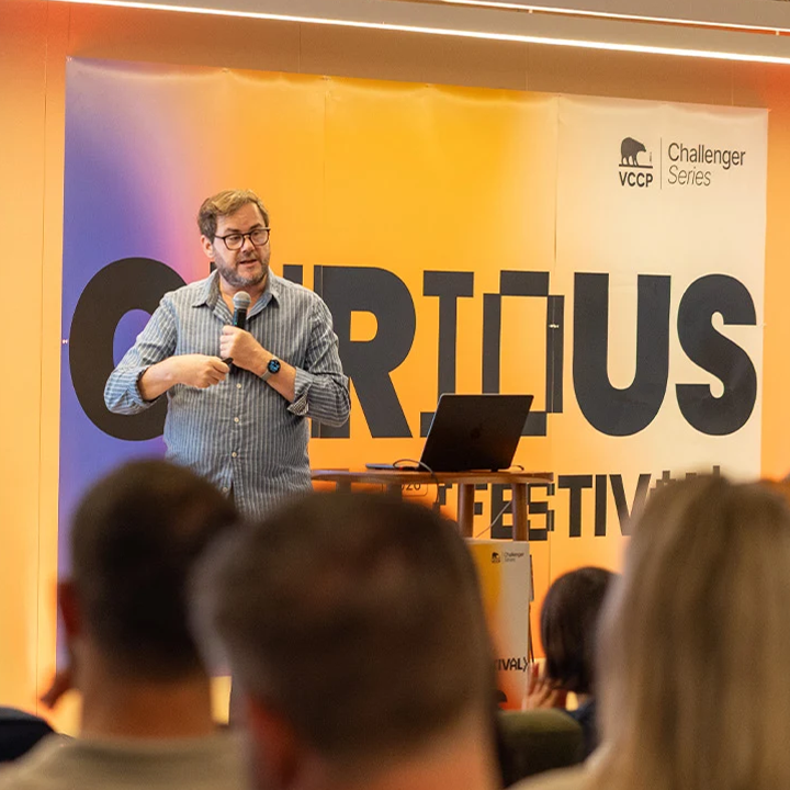 Peter Gasston presenting on stage at Curious Festival next to a screen and rows of people's head at the bottom of the image, displaying ChatGPT Intelligent UI.
