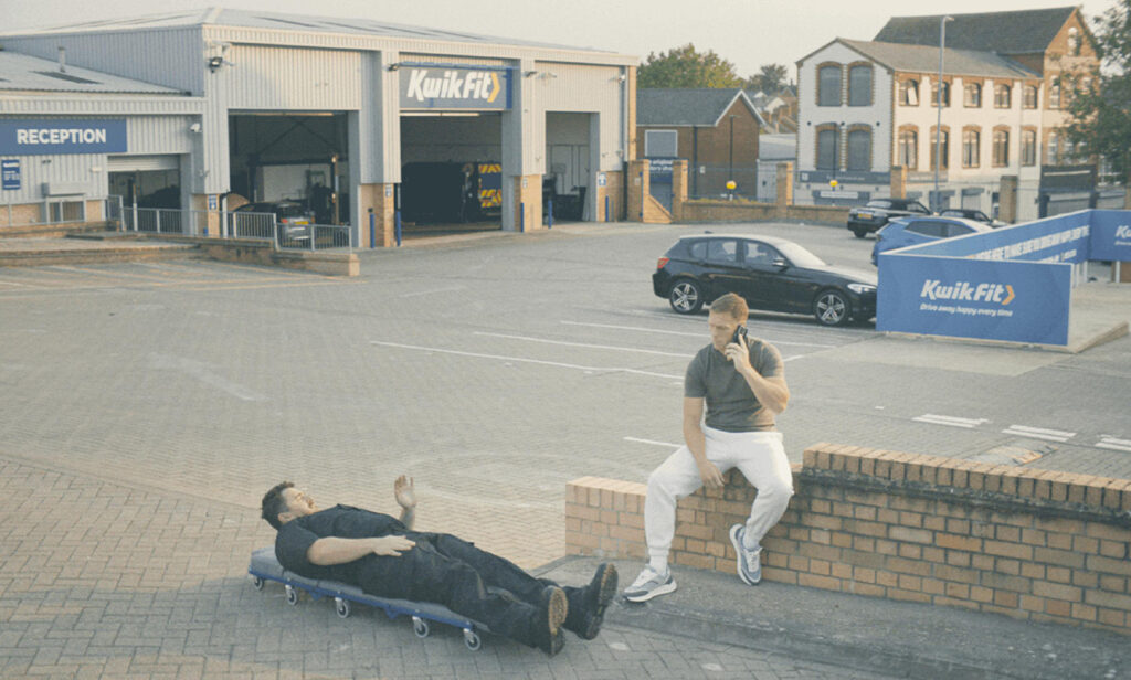 Kwik Fit mechanic lying on a creeper board outside talking to a man sitting on a brick wall using a mobile phone.