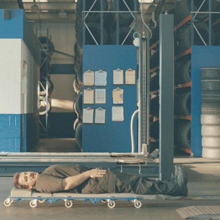 Wide banner shot of a Kwik Fit service bay with a mechanic on a creeper dolly beneath a vehicle on a hydraulic lift.