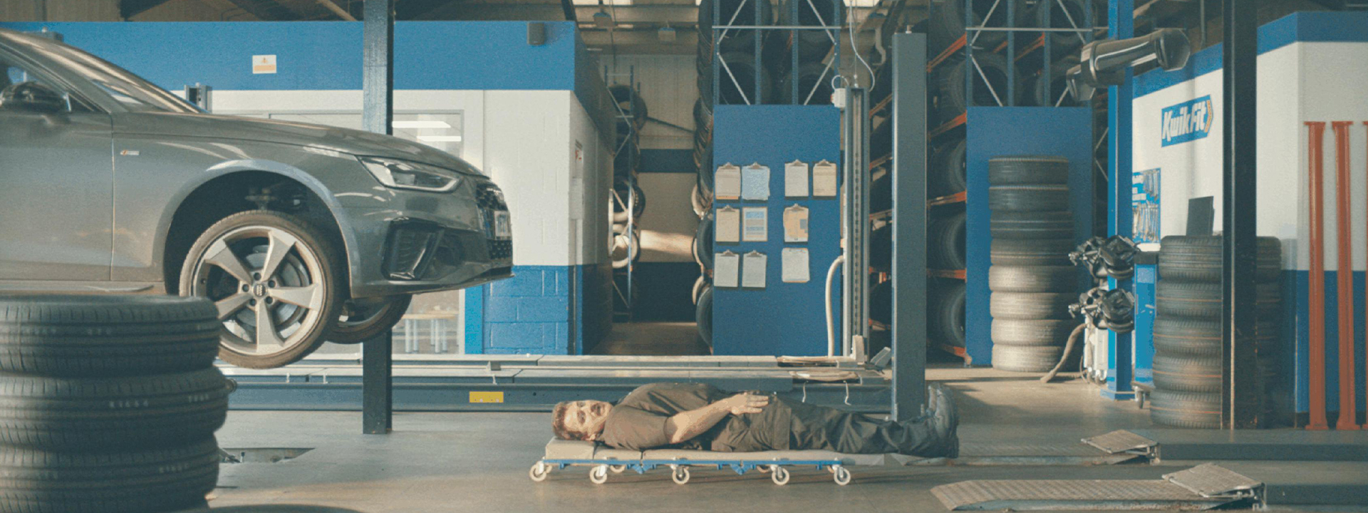 Wide banner shot of a Kwik Fit service bay with a mechanic on a creeper dolly beneath a vehicle on a hydraulic lift.