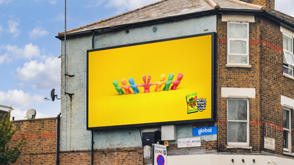 Sour Patch Kids billboard on a brick building showing candy characters playing tug of war with a red sweet splitting in half.
