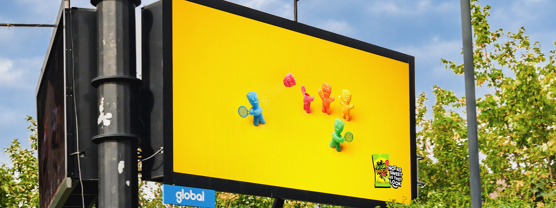 Digital billboard displaying Sour Patch Kids candy characters playing tennis using another sweet's head as a ball.