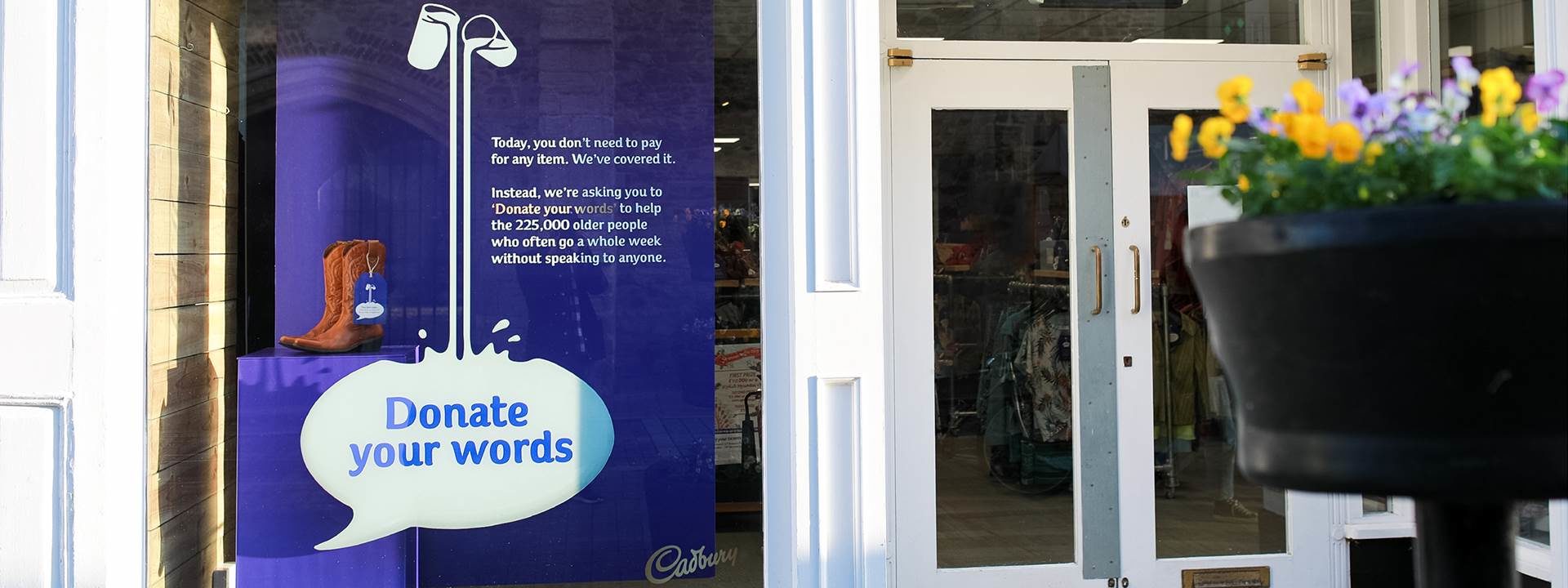 A purple Cadbury poster in a shop window. Text reads: donate your words