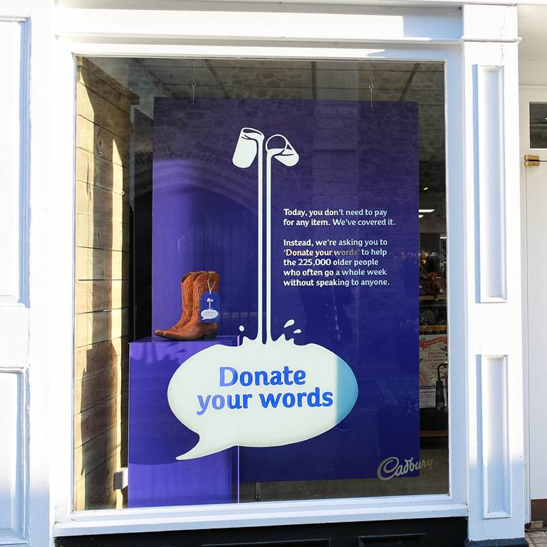 A purple Cadbury poster in a shop window. Text reads: donate your words