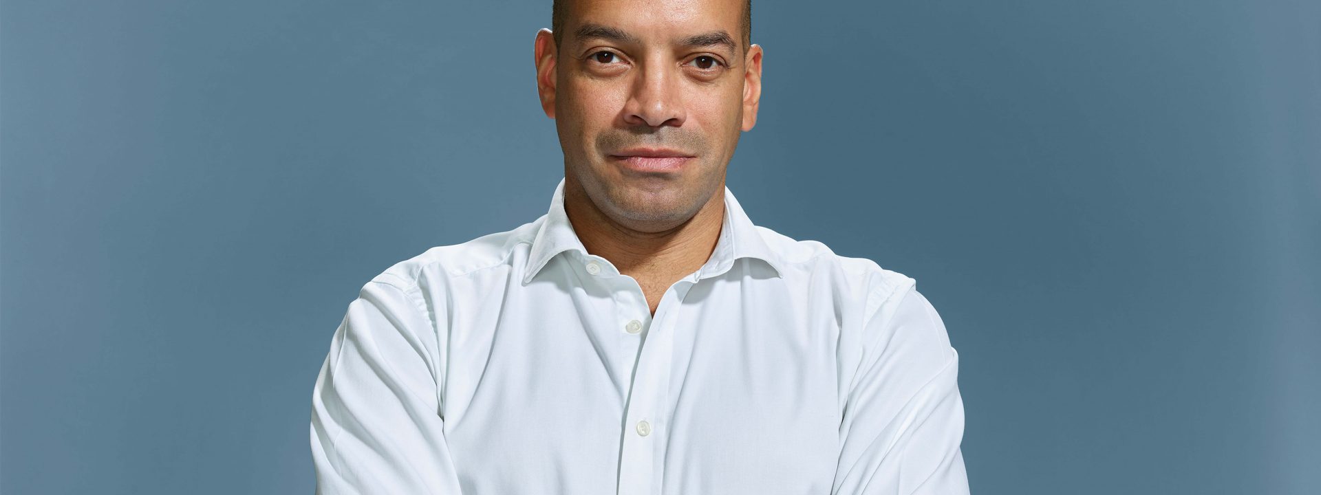 Headshot image where Dougie looks face-on at the camera, he is wearing a white shirt and has his arms crossed.