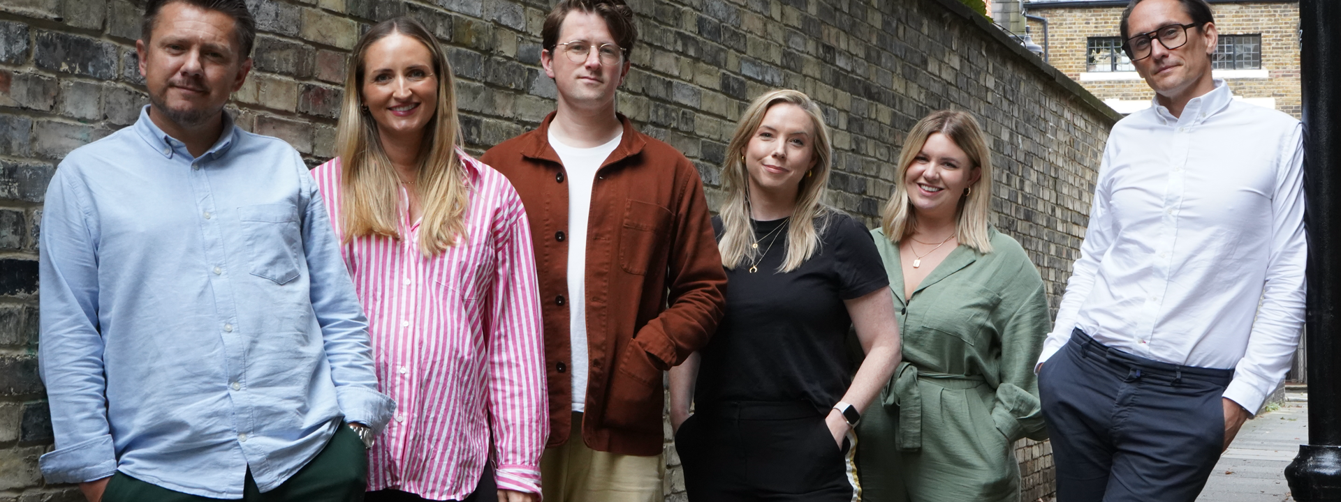 A group of six people stands in a narrow alleyway between two brick walls, posing for the photo. From left to right: 1. Simon Jenkins in a light blue shirt with rolled-up sleeves and dark green pants, standing with his hand in his pocket. 2. Becky Marsh with long blonde hair wearing a pink and white striped shirt and black pants, smiling. 3. Will Parrish with brown hair wearing glasses, a white t-shirt, and a brown jacket over beige pants. 4. Lindsay Turner with blonde hair wearing a black t-shirt and black pants, with her hands in her pockets. 5. Kim Jamieson stands with blonde hair wearing a green jumpsuit, also with her hands in her pockets. 6. James Shoreland leaning against a black lamppost. The group appears relaxed and friendly, with a backdrop of an urban brick alley and some greenery above.