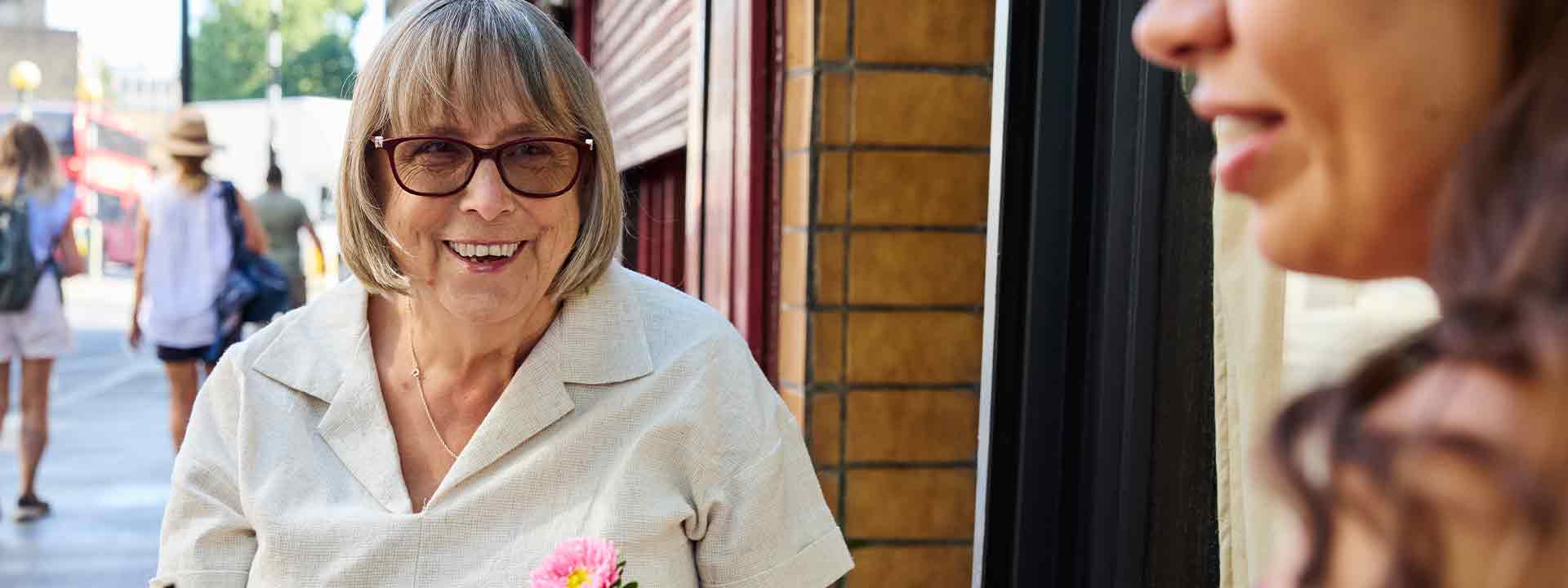 Happy senior woman with bob haircut and glasses smiling while interacting with a friend outside a shop.